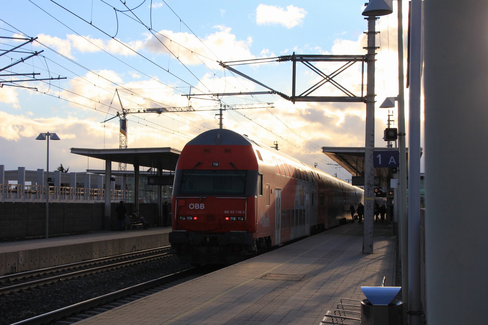 Roter Doppelstock-Wendezug der ÖBB am Bahnsteig des Bahnhofs Baden im Abendlicht, darüber Oberleitungen