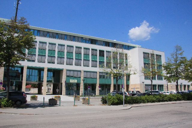 Mehrstöckiges Amtsgebäude mit weißer Fassade und grünen Fensterrahmen, davor Bäume, Blumentröge und parkende Autos, blauer Himmel