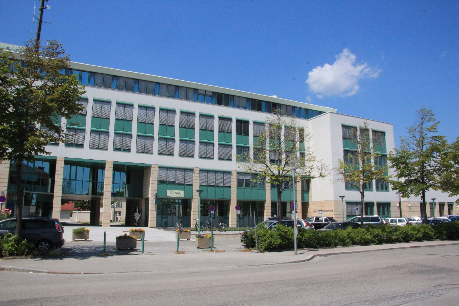 Mehrstöckiges Amtsgebäude mit weißer Fassade und grünen Fensterrahmen, davor Bäume, Blumentröge und parkende Autos, blauer Himmel