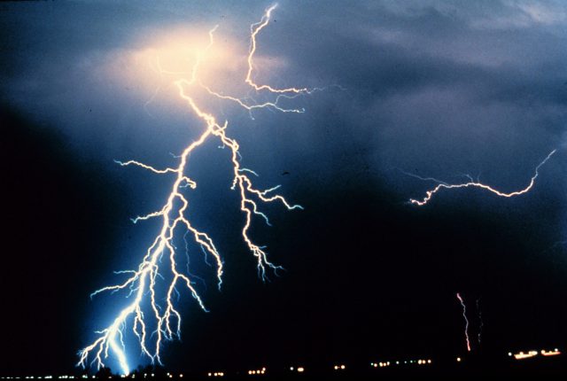 Mehrere grelle Blitze schlagen aus dunklen Gewitterwolken in den Boden, darunter Lichter in der Ferne