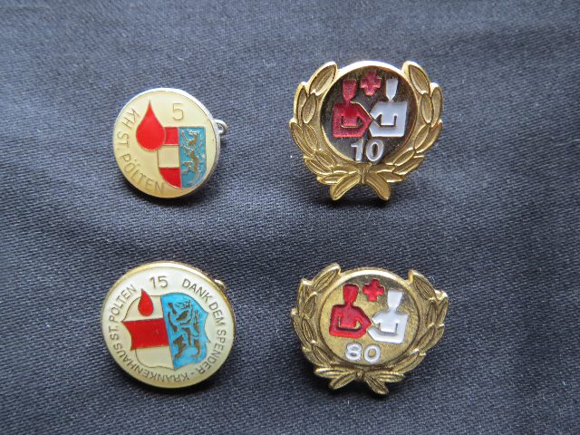 Vier Anstecknadeln für Blutspender auf dunklem Stoff, zwei mit dem Wappen von St. Pölten und Blutstropfen, zwei goldene mit Lorbeerkranz und den Zahlen 10 und 80