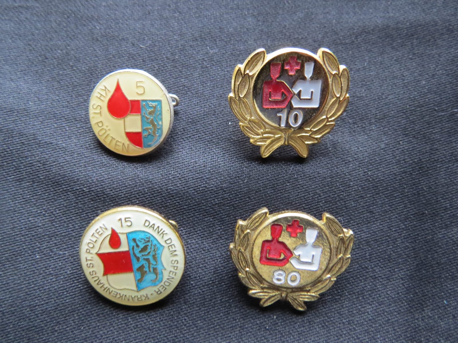 Vier Anstecknadeln für Blutspender auf dunklem Stoff, zwei mit dem Wappen von St. Pölten und Blutstropfen, zwei goldene mit Lorbeerkranz und den Zahlen 10 und 80