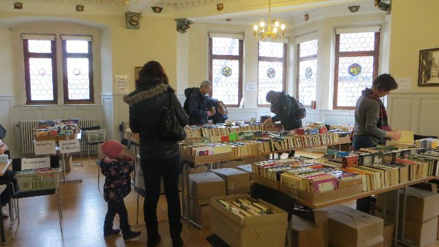 Lange Tische voller Bücher und Bücherkisten in einem hellen Saal mit Kronleuchtern, einige Menschen stöbern