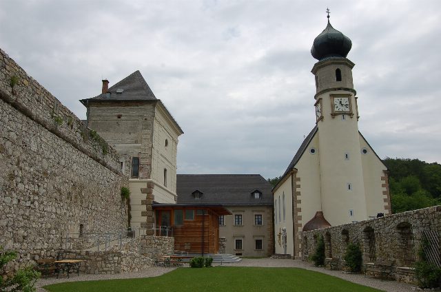 Burghof mit Rasen, links eine hohe Bruchsteinmauer und ein Wohnturm, rechts eine helle Kirche mit Zwiebelturm und Uhr