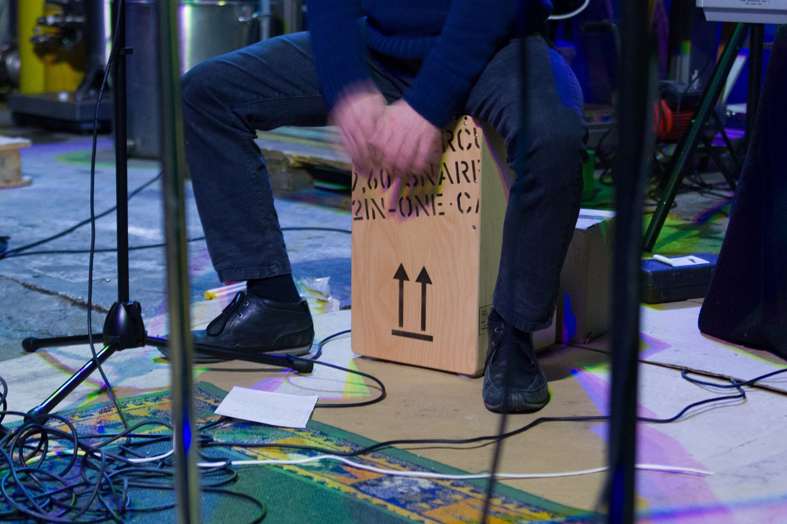 Symbolbild: Ein Musiker sitzt auf einer Holzkiste, einem Cajón, und schlägt mit den Händen darauf, rundherum Mikrofonständer und Kabel