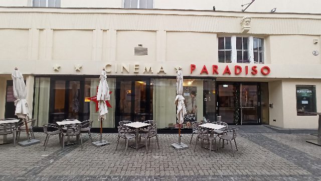 Eingang des Cinema Paradiso in der Beethovengasse in Baden mit rotem Schriftzug, davor Tische und zusammengeklappte Sonnenschirme des Schanigartens