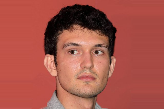 Porträt eines jungen Mannes mit dunklem, lockigem Haar und grauem Jeanshemd vor rotem Hintergrund