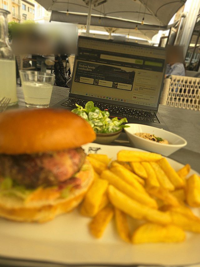 Burger mit Pommes auf einem Teller im Schanigarten des El Gaucho Baden, dahinter ein Glas Soda Zitrone und ein Laptop mit der Startseite von badenliefert.at