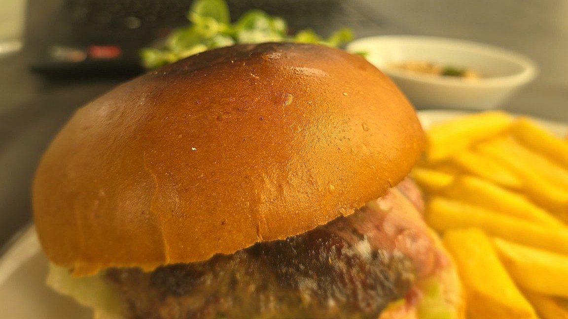 Burger mit glänzendem Brötchen, Fleischlaibchen und Salat, daneben Pommes, dahinter eine Schale grüner Salat und ein Laptop – Mittagsmenü im El Gaucho Baden