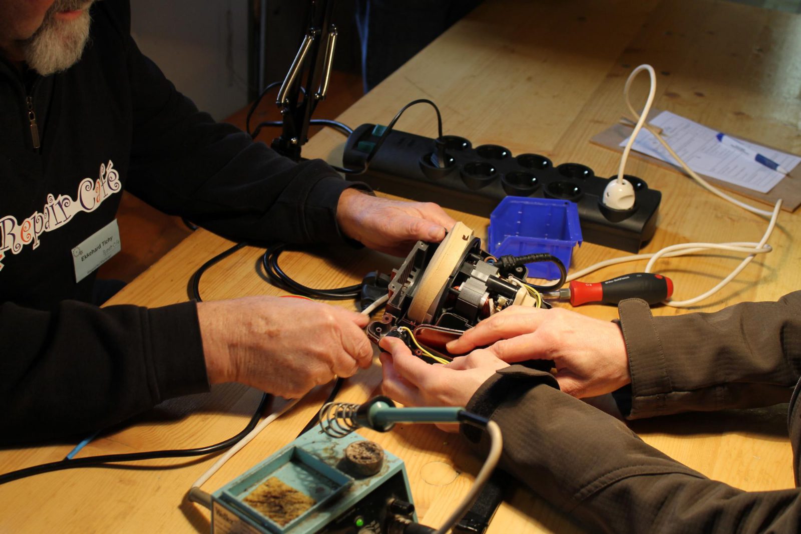 Zwei Paar Hände halten ein geöffnetes Elektrogerät über einem Holztisch, daneben Steckdosenleiste, Schraubenzieher und Lötkolben