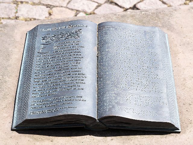 Ein aufgeschlagenes Buch aus Metall auf einem Steinsockel, links der Titel Emil und die Detektive mit einer Textstelle, rechts derselbe Text in Blindenschrift