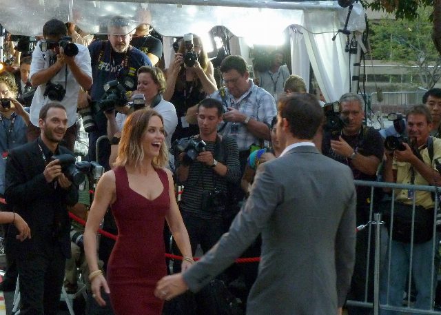 Emily Blunt im roten Kleid und John Krasinski von hinten auf einem roten Teppich, dahinter eine Reihe von Fotografen mit Kameras