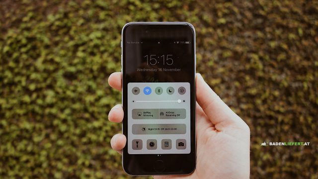 Hand hält ein Smartphone vor einer grünen Hecke, auf dem Bildschirm steht oben links „No Service“, darunter das Kontrollzentrum mit WLAN-Symbol