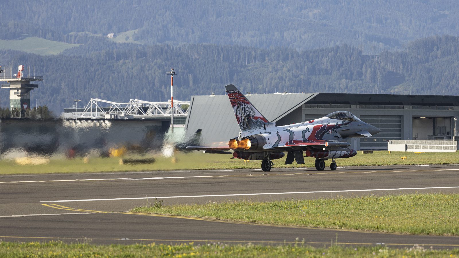 Kampfjet in grau-roter Sonderlackierung mit Tigerkopf am Leitwerk beim Start auf einer Piste, aus den Triebwerken leuchtet Nachbrennerfeuer, im Hintergrund Hangar und bewaldete Hügel
