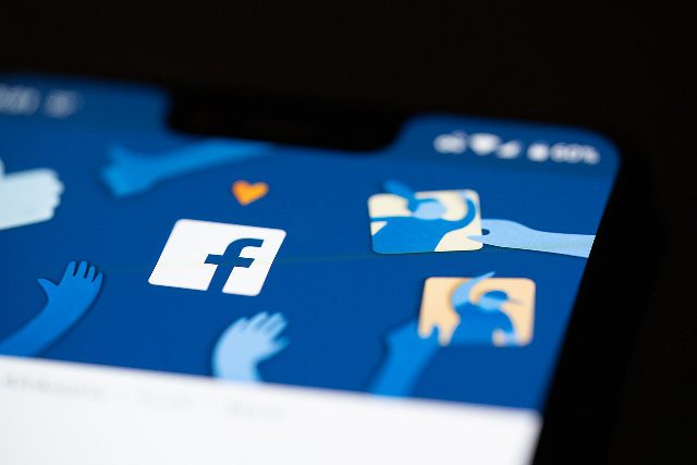 Bildschirm eines Smartphones mit dem Startbild der Facebook-App: weißes F-Logo auf blauem Grund zwischen gezeichneten Händen