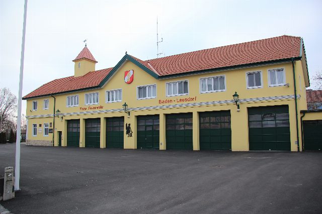Feuerwehrhaus Baden-Leesdorf: gelber Bau mit rotem Dach, Türmchen, Stadtwappen im Giebel und sieben grünen Toren