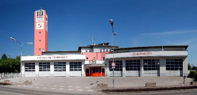 Feuerwehrhaus der Freiwilligen Feuerwehr Leobersdorf mit Fahrzeughallen und dem rosa Feuerwehrturm links, davor der leere Vorplatz