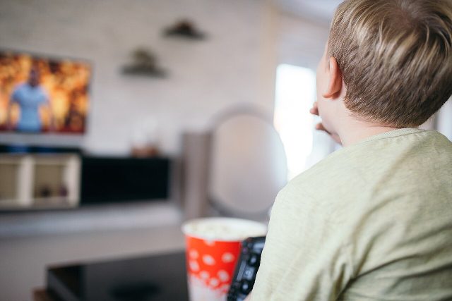Ein Bub sitzt mit Popcornbecher und Fernbedienung im Wohnzimmer und schaut auf den Fernseher, von hinten fotografiert