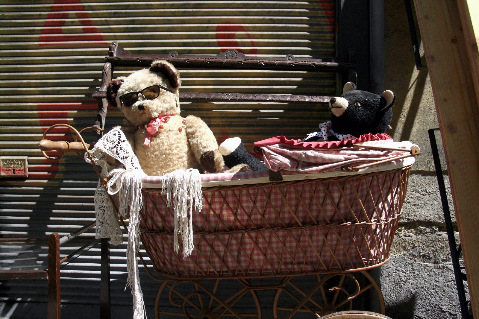 Zwei alte Teddybären, einer mit Sonnenbrille, sitzen in einem geflochtenen Puppenwagen vor einem Rollladen