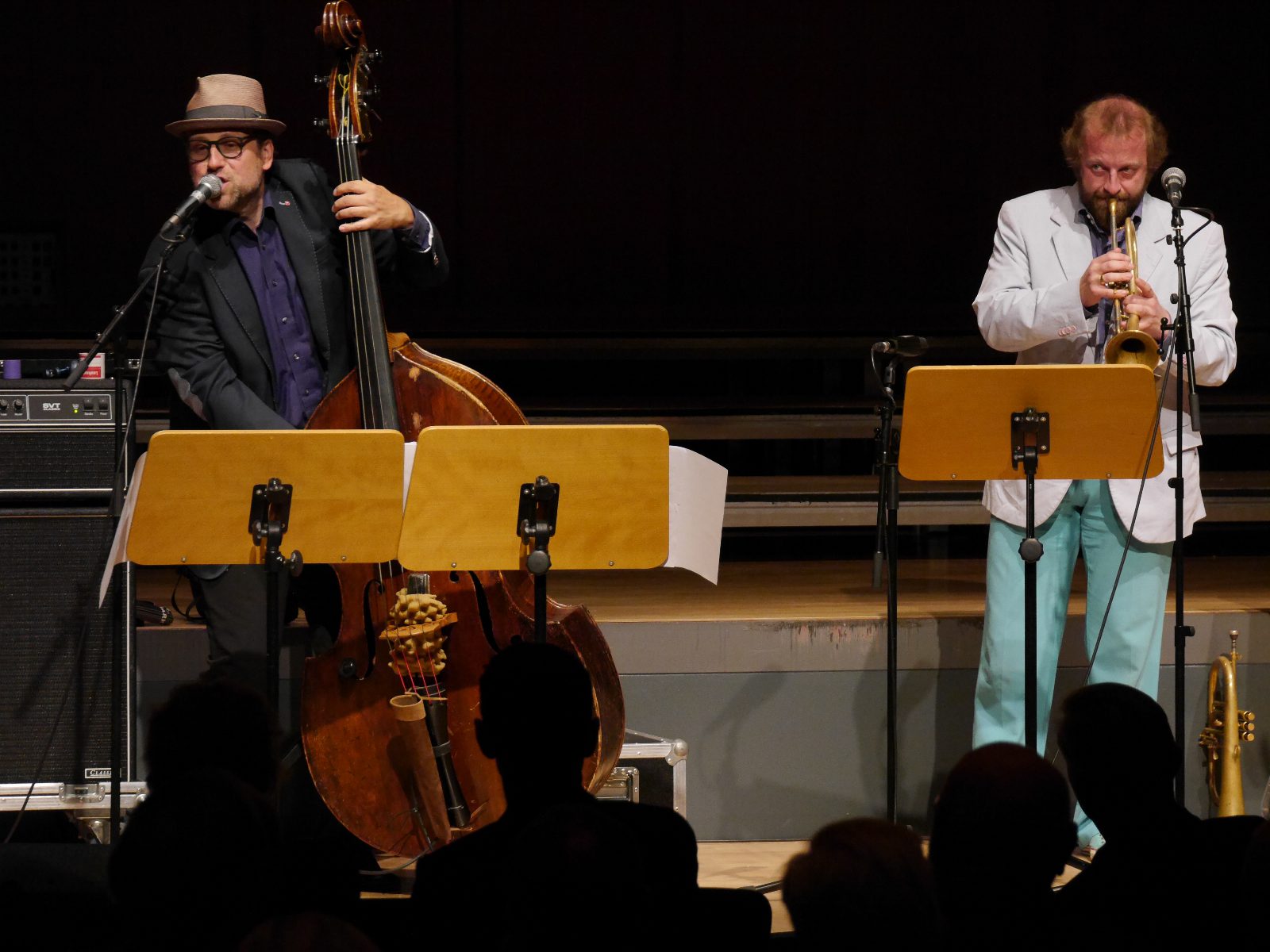 Ein Musiker mit Hut singt am Kontrabass, rechts spielt ein Musiker im hellen Anzug Trompete, vor beiden Notenständer, im Vordergrund Köpfe des Publikums