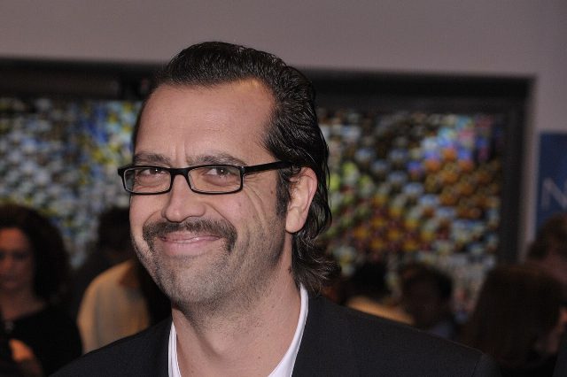 Der Kabarettist Gerald Fleischhacker mit dunkler Brille, Schnurrbart und zurückgekämmtem Haar lächelt bei einer Veranstaltung in die Kamera