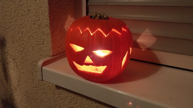 Geschnitzter Kürbis mit Zackenmuster, schrägen Augen und breitem Grinsen, von einer Kerze innen beleuchtet, auf einem Fensterbrett