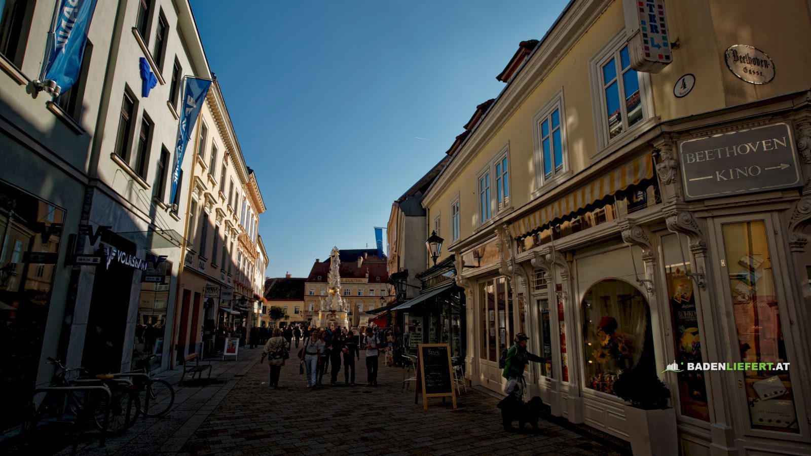 Fußgängerzone in Baden mit Geschäften links und rechts, Spaziergänger auf dem Pflaster und im Hintergrund die Pestsäule am Hauptplatz