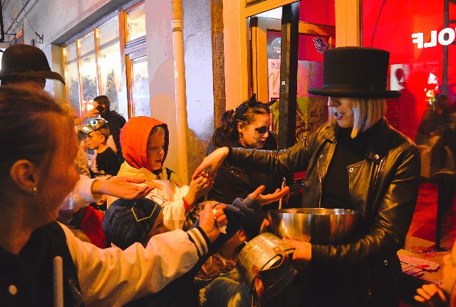 Halloween in Bielsko-Biała in Polen: Eine Frau mit Zylinder verteilt aus einer Metallschüssel Süßigkeiten an Kinder in Kapuzen und Masken