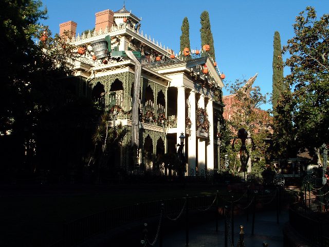 Weißes Herrenhaus mit Säulen und verzierten Balkonen, auf dem Dach und am Geländer orange Kürbisse, davor dunkle Bäume und ein Kettenzaun