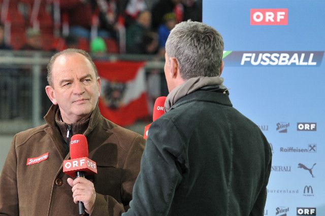 Herbert Prohaska in brauner Jacke mit ORF-Mikrofon im Interview, im Hintergrund eine Fußballtribüne und eine Sponsorwand