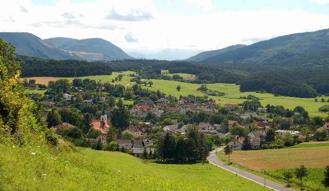 Blick von einem Wiesenhang auf Hernstein mit Kirchturm, Häusern und bewaldeten Hügeln im Hintergrund