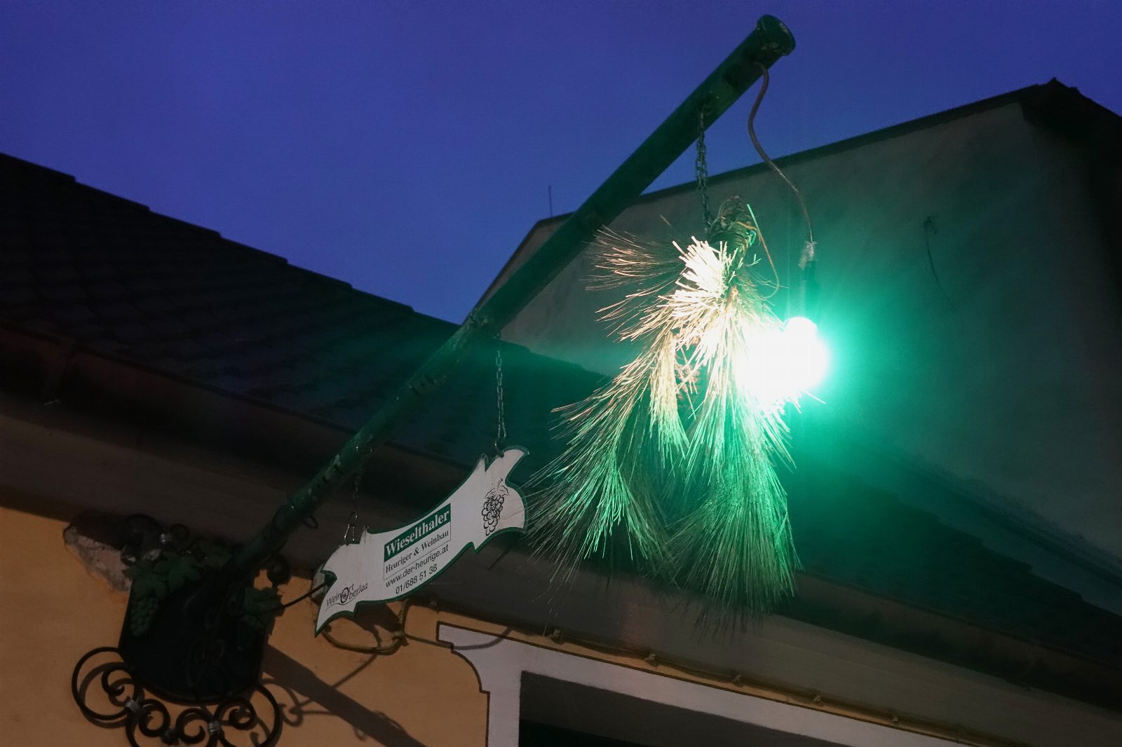 Grün beleuchteter Föhrenbuschen an einer Stange über einem Hauseingang in der Abenddämmerung, darunter ein kleines Schild