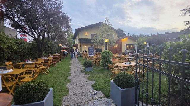 Gastgarten in der Abenddämmerung: gelbe Holztische und Klappsessel auf der Wiese zu beiden Seiten eines Plattenwegs, dahinter ein gelbes Haus mit brennenden Laternen, vorne ein schmiedeeisernes Gartentor und Buchskugeln in grauen Trögen