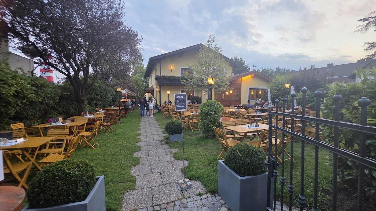 Gastgarten in der Abenddämmerung: gelbe Holztische und Klappsessel auf der Wiese zu beiden Seiten eines Plattenwegs, dahinter ein gelbes Haus mit brennenden Laternen, vorne ein schmiedeeisernes Gartentor und Buchskugeln in grauen Trögen