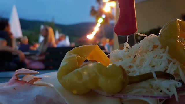 Belegtes Brot mit Schinken, Käse und Pfefferoni, dahinter ein Heurigengarten am Abend mit Lichterkette