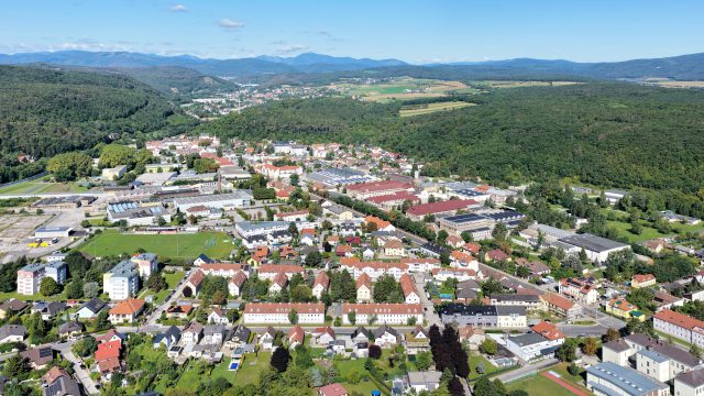 Luftbild von Hirtenberg von Südosten: Wohnhäuser, Sportplatz und Gewerbebauten im Tal, dahinter bewaldete Hügel und die Berge