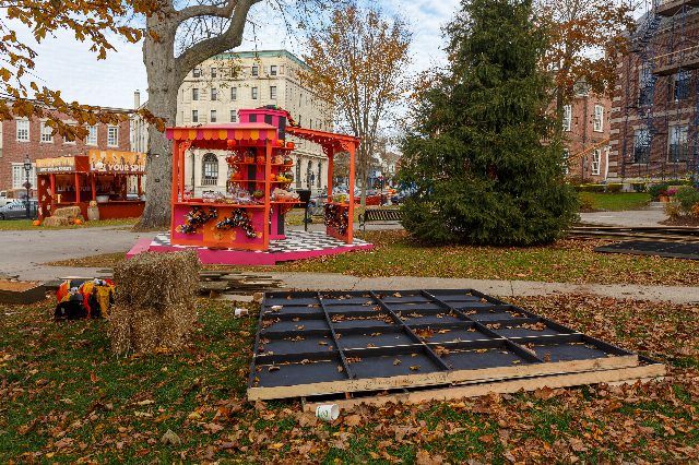Ein pinkfarbener Jahrmarktstand mit Süßigkeiten auf einer Wiese voller Herbstlaub, davor Strohballen und ein am Boden liegendes Kulissenteil, im Hintergrund Bäume und Backsteinhäuser