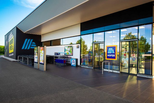 Eingangsbereich einer Hofer-Filiale mit Glasfront, Hofer-Logo, Einkaufswagen und zwei Werbesäulen