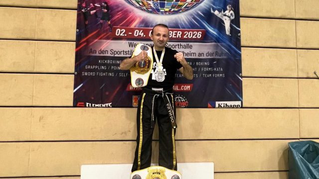 Mikail Cetiner steht mit Meistergürtel und Medaille vor dem Banner der WFMC-Weltmeisterschaft, auf dem Termin und Austragungsort in Mülheim an der Ruhr stehen