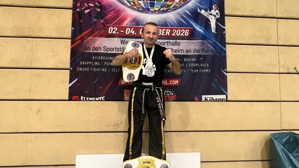 Mikail Cetiner steht mit Meistergürtel und Medaille vor dem Banner der WFMC-Weltmeisterschaft, auf dem Termin und Austragungsort in Mülheim an der Ruhr stehen