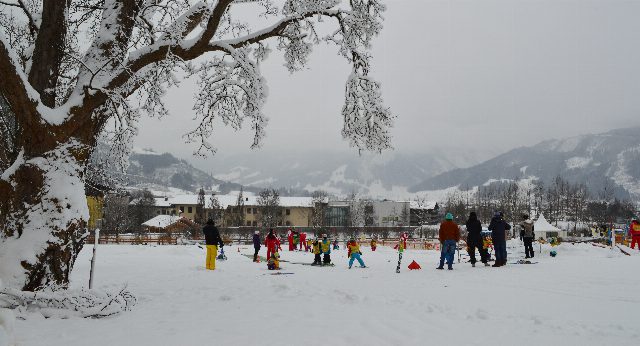 Verschneite Übungswiese mit kleinen Kindern auf Ski in bunten Anzügen, Erwachsene stehen daneben, links ein verschneiter Baum, hinten Berge im Nebel