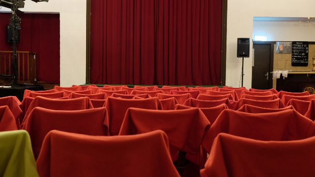 Kleiner Theatersaal mit geschlossenem rotem Vorhang, davor Stuhlreihen mit roten Hussen, rechts ein Lautsprecher und eine Schank