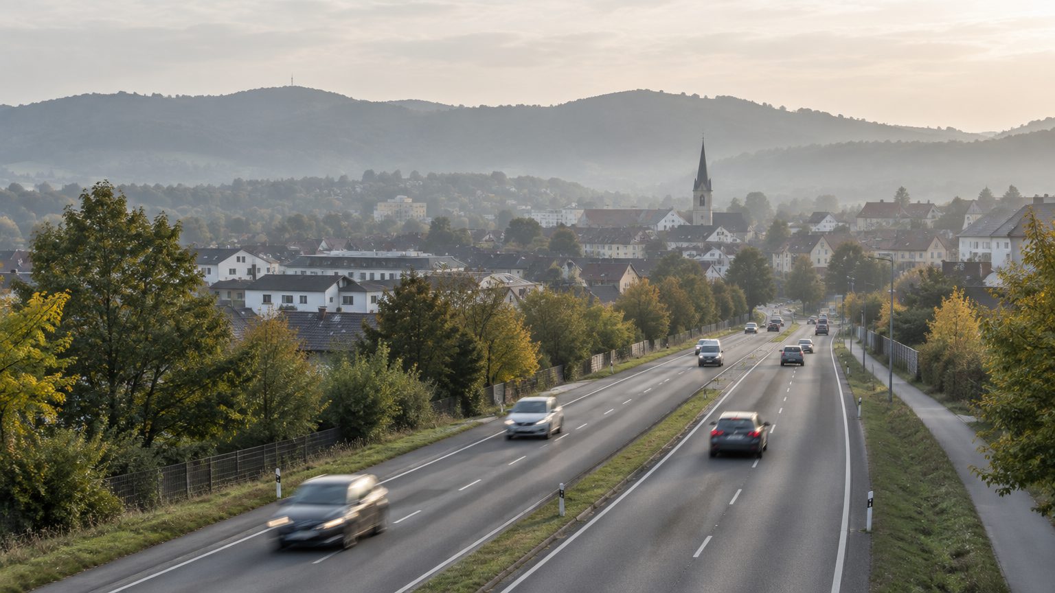 Symbolbild: eine stark befahrene Einfallstraße am Ortsrand im Morgenlicht, dahinter Dächer einer Kleinstadt