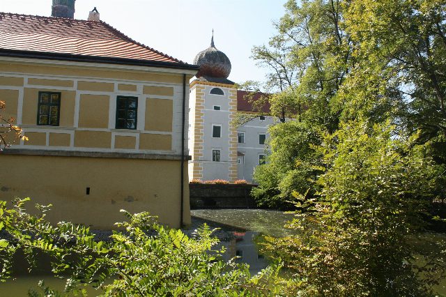 Gelbes Schlossgebäude am Wassergraben, dahinter ein heller Turm mit Zwiebelhaube zwischen Bäumen