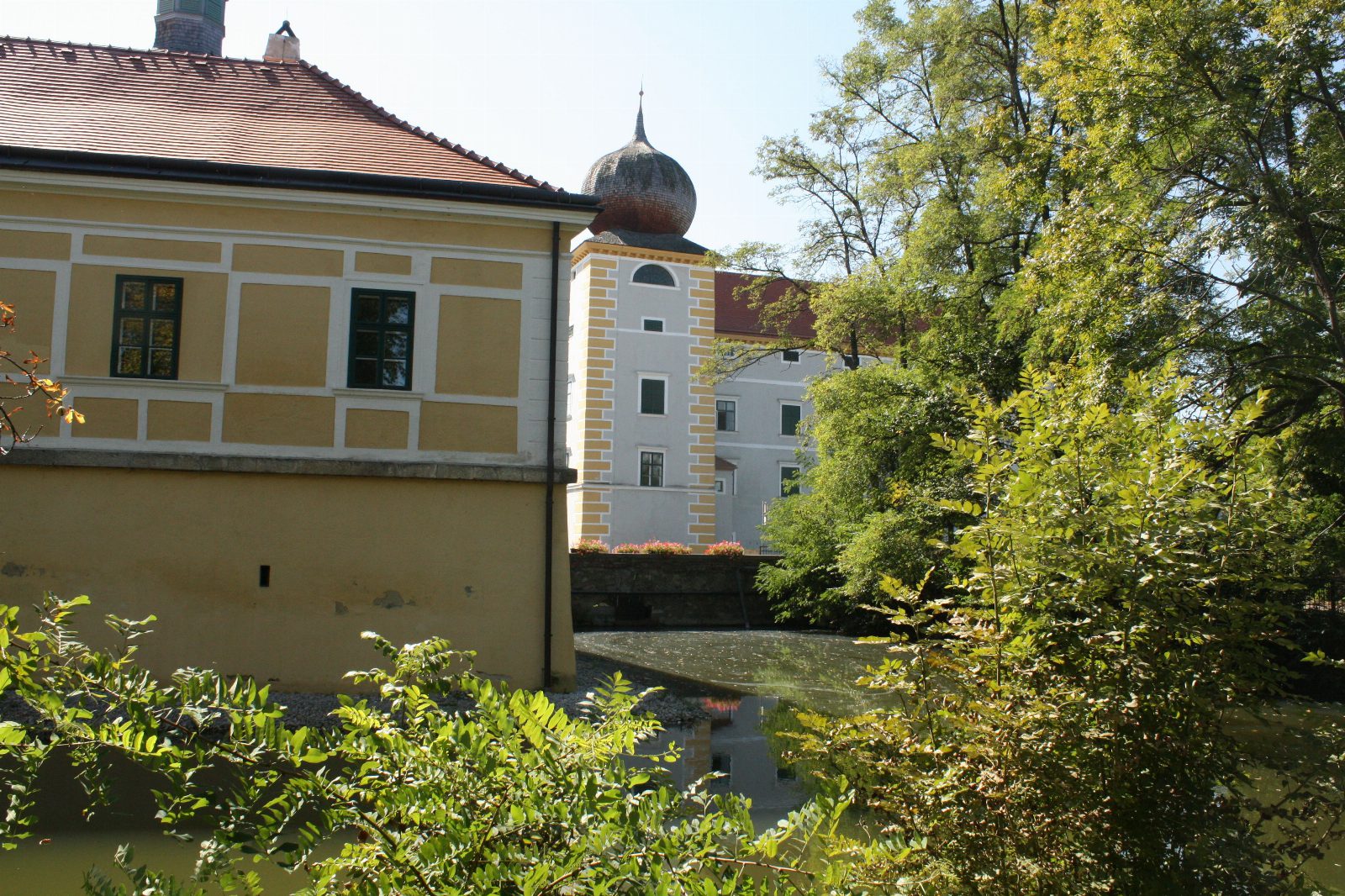 Gelbes Schlossgebäude am Wassergraben, dahinter ein heller Turm mit Zwiebelhaube zwischen Bäumen