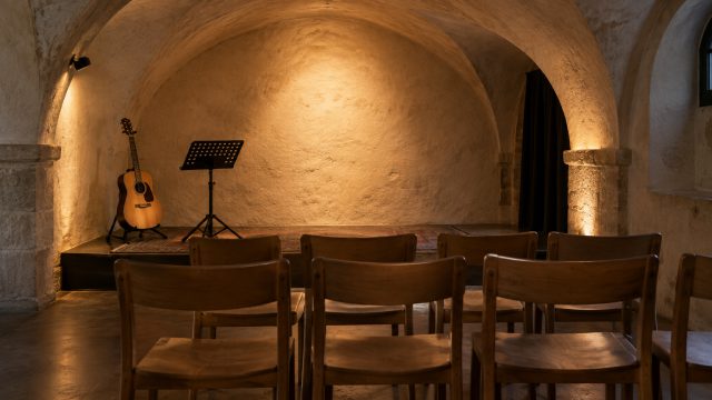 Symbolbild: ein kleiner Saal in einem alten Gewölbe vor Beginn, Reihen schlichter Holzstühle, am Rand eine akustische Gitarre auf einem Ständer und ein Notenpult