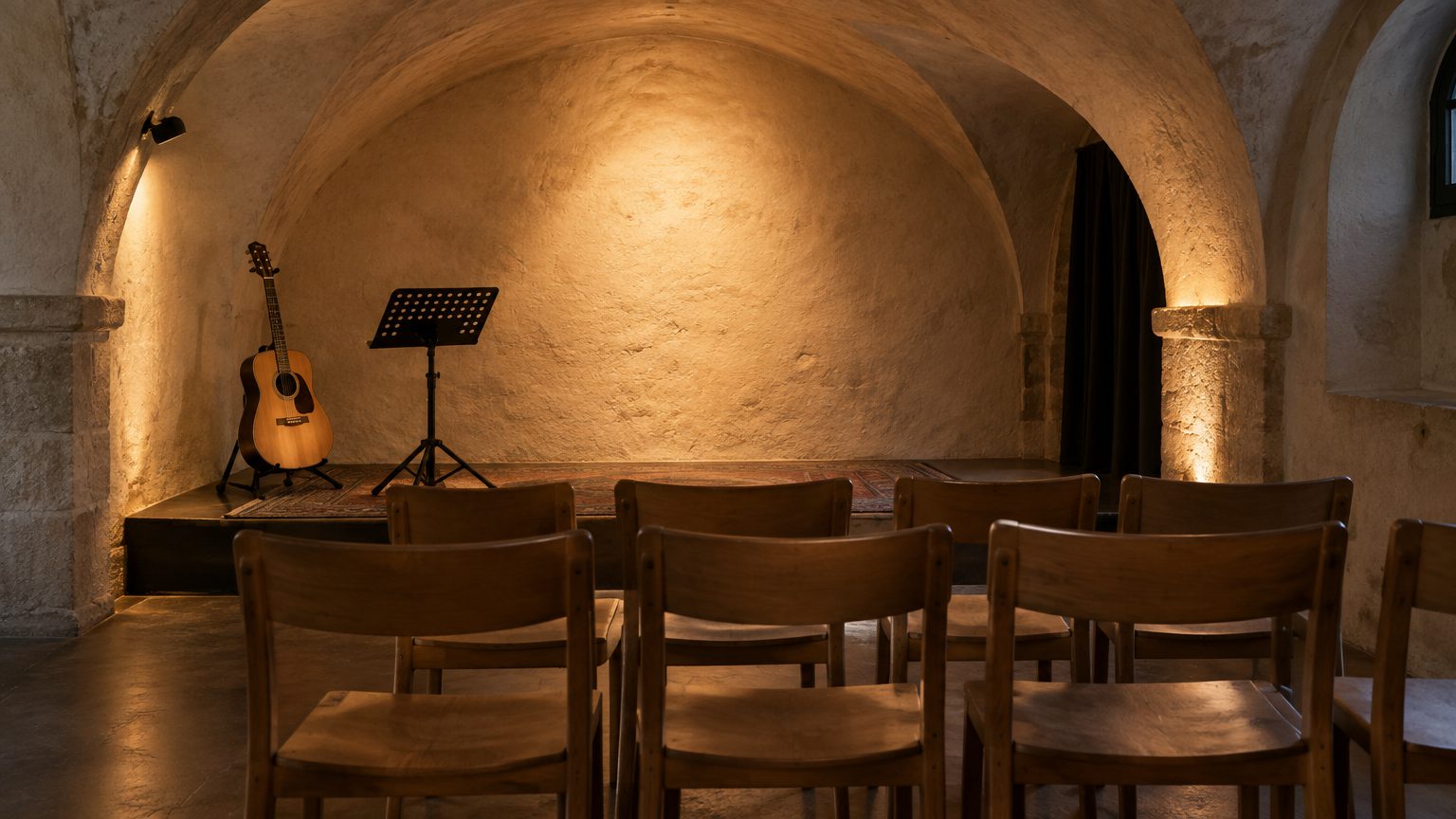 Symbolbild: ein kleiner Saal in einem alten Gewölbe vor Beginn, Reihen schlichter Holzstühle, am Rand eine akustische Gitarre auf einem Ständer und ein Notenpult