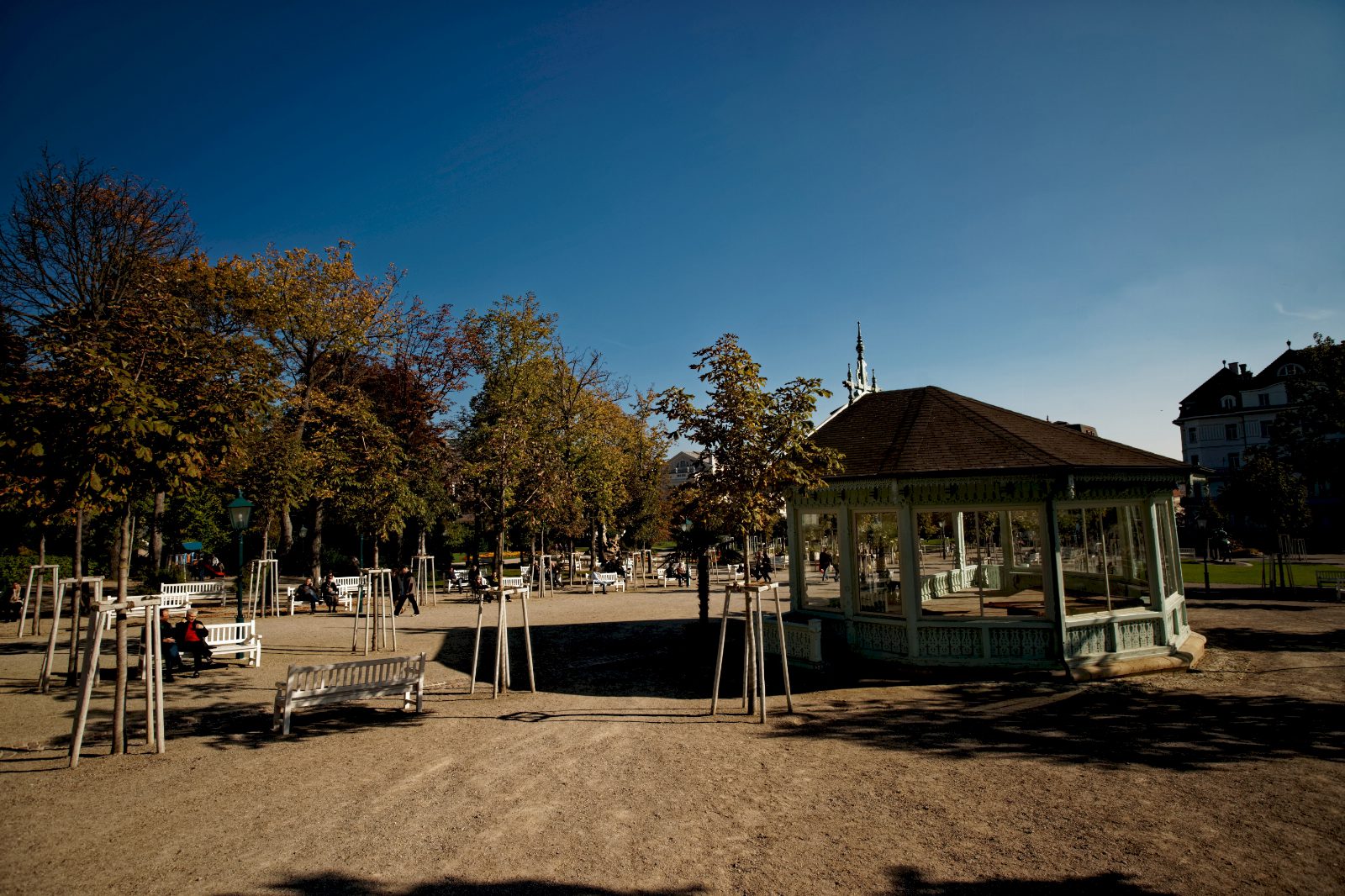 Herbstlich gefärbte Bäume und ein achteckiger Pavillon mit Holzdach auf einem sonnigen Kiesplatz im Kurpark Baden, daneben Parkbänke