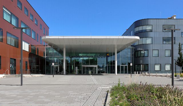 Haupteingang des Landesklinikums Baden mit weißem Vordach zwischen einem rotbraunen und einem grauen Gebäudeteil, davor ein gepflasterter Vorplatz