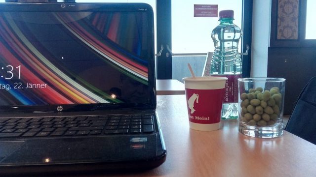 Aufgeklappter schwarzer HP-Laptop auf einem Holztisch, daneben ein Kaffeebecher, eine Wasserflasche und ein Glas mit grünen Nüssen